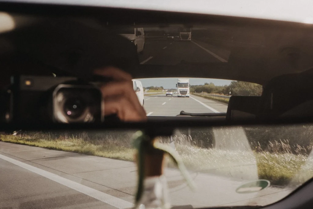 Blick durch den Rückspiegel auf die Autobahn, im Vordergrund eine Hand am Lenkrad