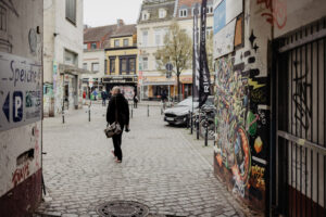 Andreas geht mit einer Tasche eine Kopfsteinpflasterstraße entlang, im Hintergrund bunte Altstadthäuser und ein Wandgemälde