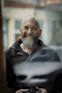 Porträtfoto von Andreas, lächelnd hinter einer Fensterscheibe mit Spiegelungen